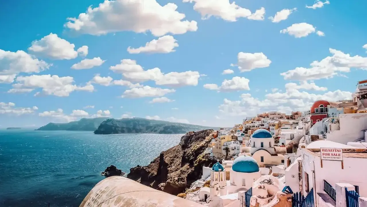 Santorini, Grecja. Białe domy z niebieskimi kopułami kościołów na zboczu góry, nad turkusowym morzem. Marzysz o podróży? Sprawdź, ile się leci do Grecji!