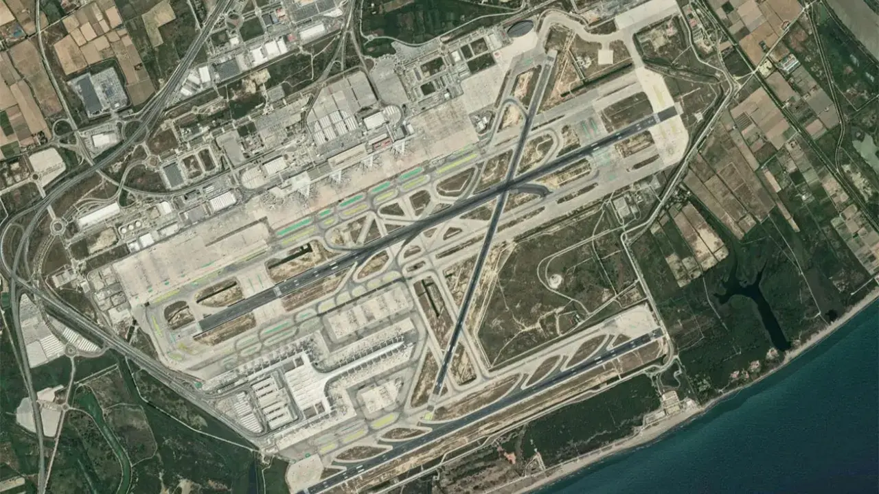 Widok satelitarny lotniska Barcelona El Prat. Widać pasy startowe, drogi kołowania, terminale i otaczające tereny rolnicze oraz morze.