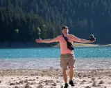 Mężczyzna z aparatem stoi na kamienistej plaży nad turkusowym jeziorem, zalesione wzgórza w tle.