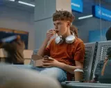 Chłopak w słuchawkach je w poczekalni lotniska. Zastanawia się, od ilu lat można latać samemu.
