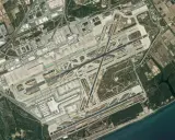 Widok satelitarny lotniska Barcelona El Prat. Widać pasy startowe, drogi kołowania, terminale i otaczające tereny rolnicze oraz morze.