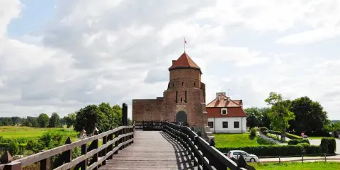 Drewniany most prowadzi do ceglanego zamku z czerwoną wieżą i białym budynkiem.