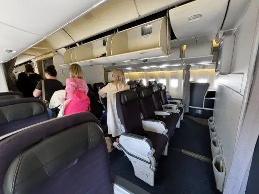 Wnętrze samolotu z rzędami foteli i pasażerami w przejściu.
