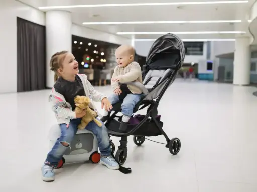 Dwie dziewczynki, jedna siedzi na walizce z misiem, druga w wózku, śmieją się w jasnym wnętrzu.