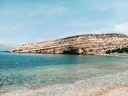 Plaża z kamienistym brzegiem i skalistym klifem w tle, nad turkusowym morzem.