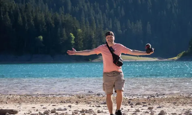 Mężczyzna z aparatem stoi na kamienistej plaży nad turkusowym jeziorem, zalesione wzgórza w tle.