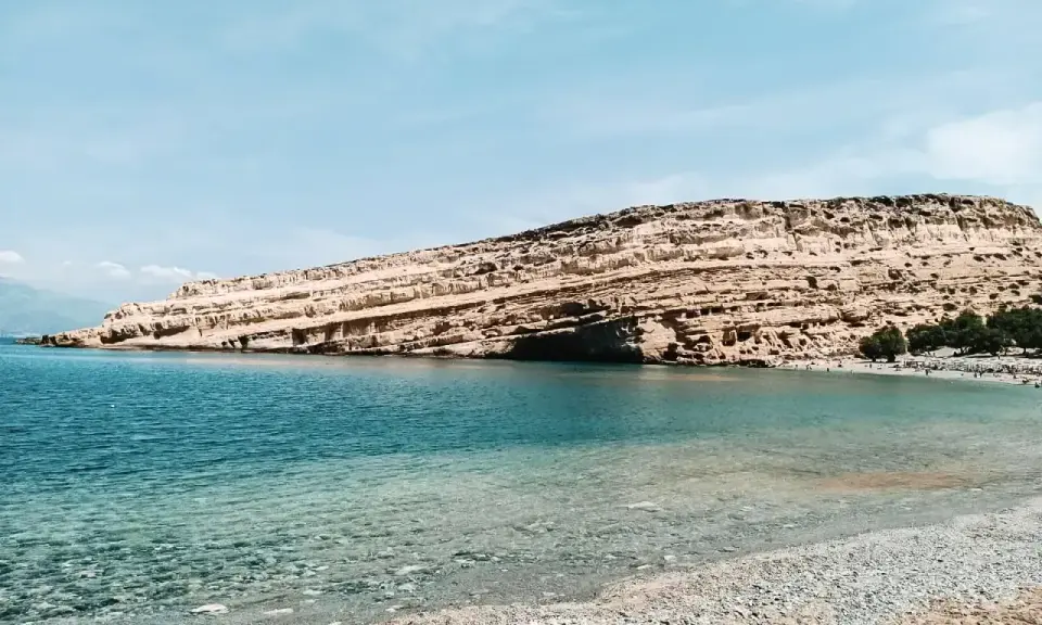 Plaża z kamienistym brzegiem i skalistym klifem w tle, nad turkusowym morzem.
