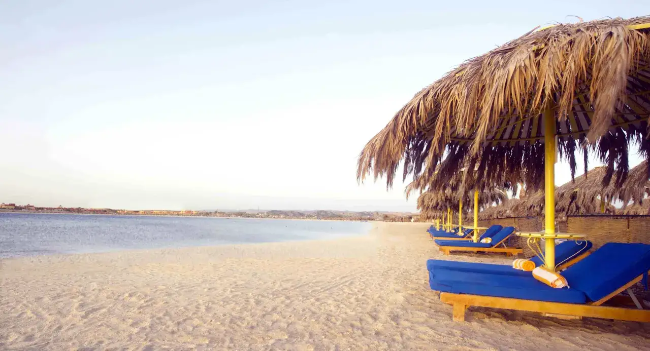 Plaża z leżakami i parasolami z liści palmowych. W tle widać morze i zabudowania, idealne miejsce na wakacje, zgodnie z hilton marsa alam nubian resort opinie.