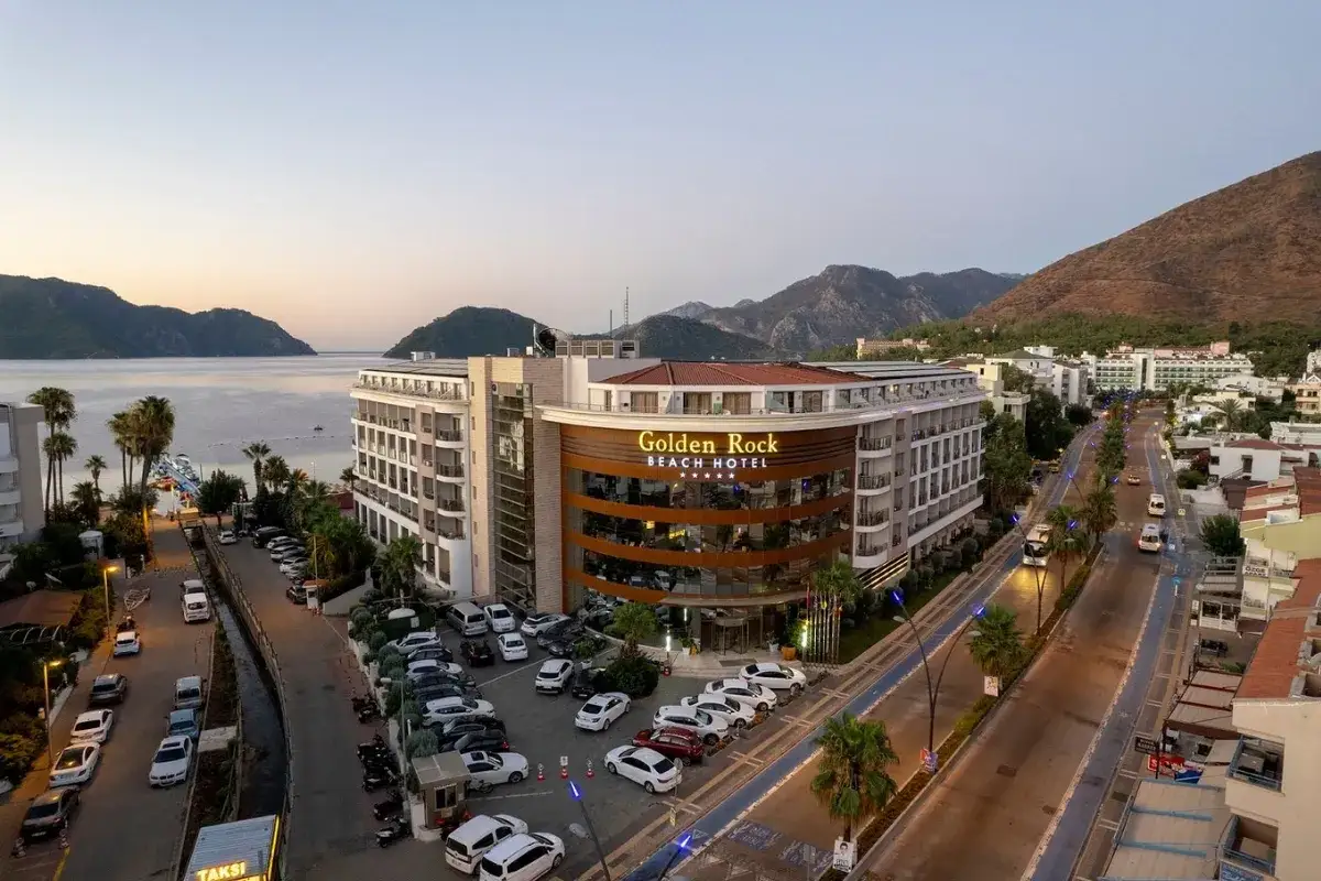 Widok na hotel Golden Rock Beach Hotel z parkingiem i drogą. W tle morze i góry.