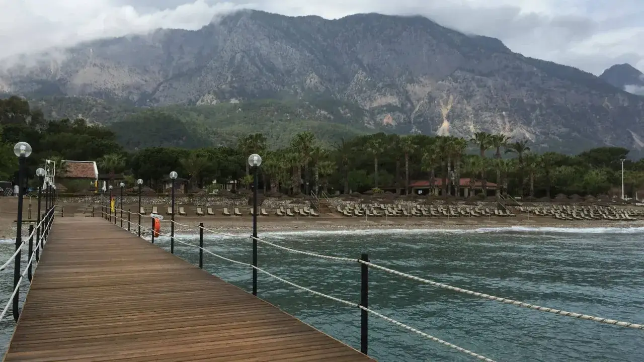 Molo prowadzi do plaży z leżakami i palmami, w tle góry. Idealne miejsce na wakacje, jak sugerują champion holiday village opinie.