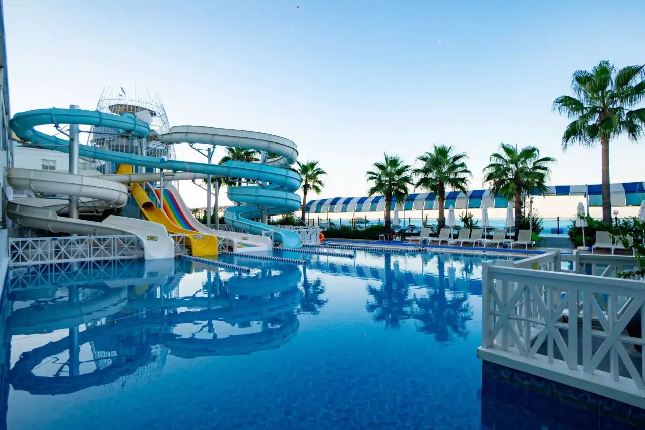 Basen z wodnymi zjeżdżalniami w Orange County Resort Alanya. Palmy i leżaki przy błękitnej wodzie.