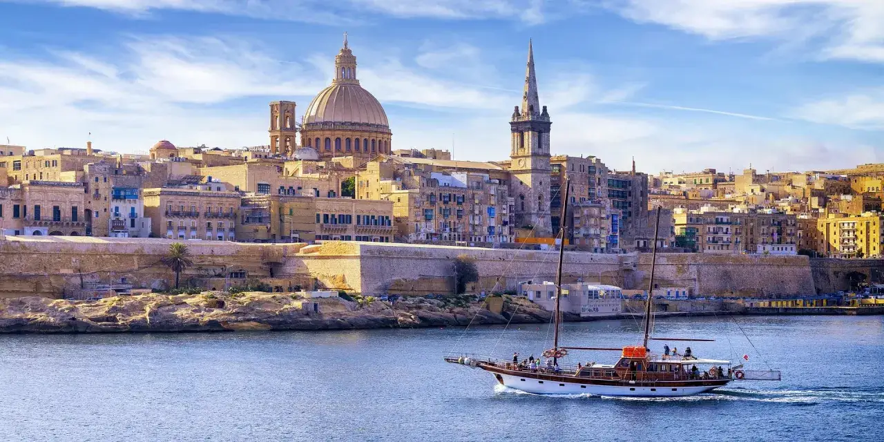 Malta, z żaglowcem na pierwszym planie i kopułą katedry w tle. Czas zacząć planować, ile się leci na Maltę!